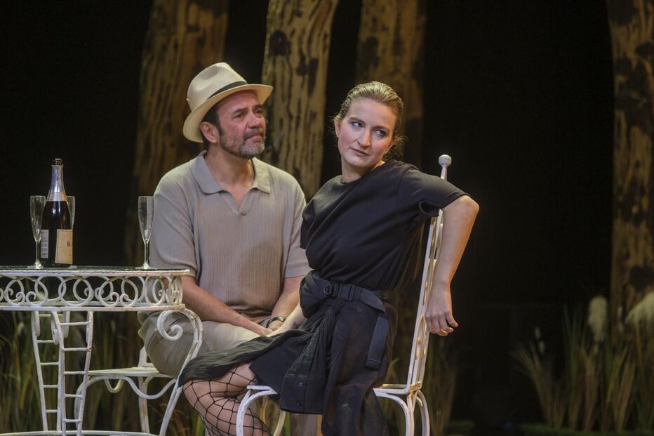 Espetáculo teatral com dois atores, mesa e champanhe em palco.
