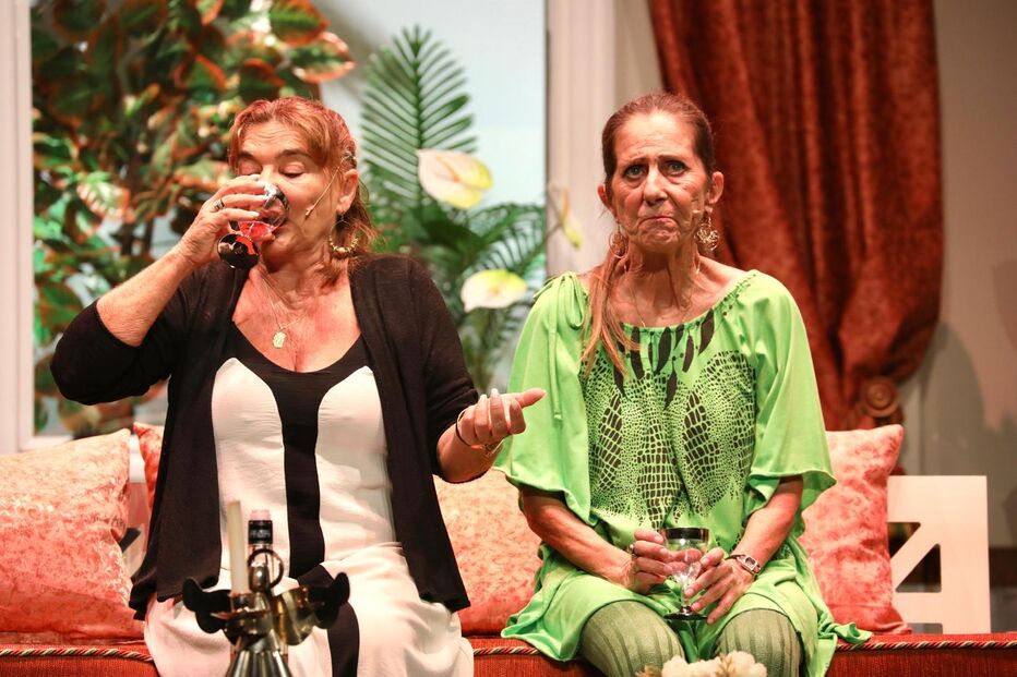 Duas mulheres num palco, uma bebe, a outra parece incomodada