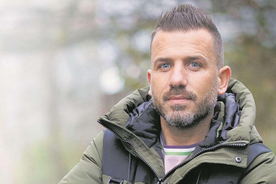 Homem de braços cruzados com casaco verde e preto num ambiente natural