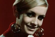 Twiggy sorri com grandes brincos dourados e um penteado elegante