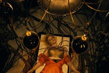 Mulher deitada, ligada a cabos, num cenário que evoca ficção científica ou fantasia