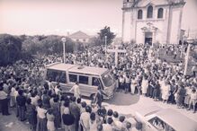 Multidão reúne-se para um funeral, com carro funerário e igreja visíveis