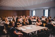 Pessoas assistem a uma apresentação numa sala com mesas e cadeiras