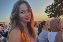 Mulher sorri num evento ao ar livre, com pessoas e um pôr do sol ao fundo