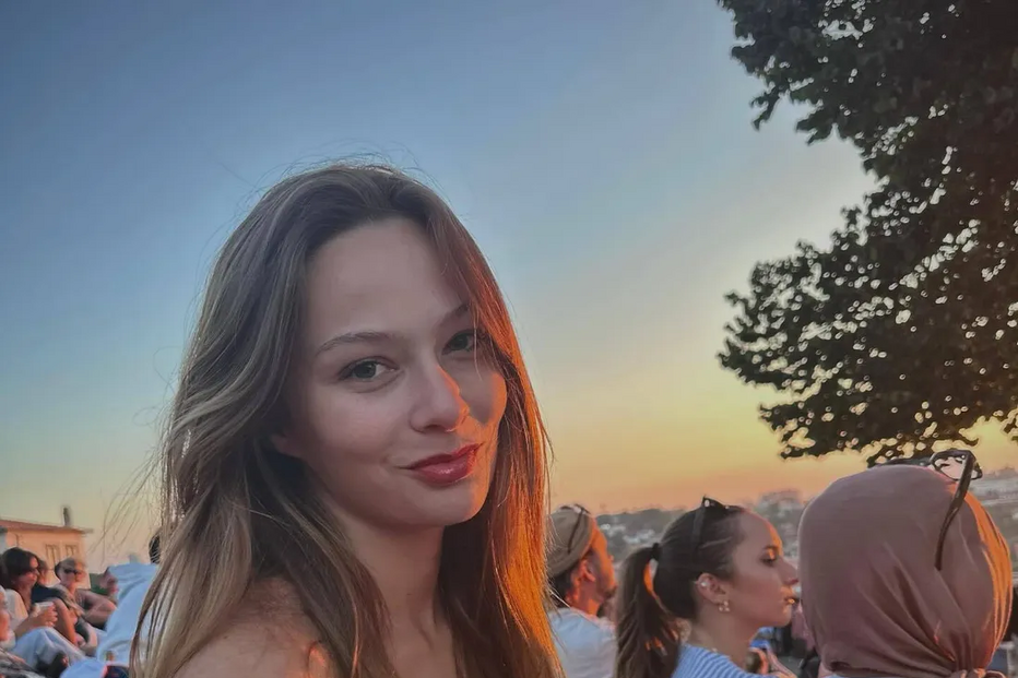 Mulher sorri num evento ao ar livre, com pessoas e um pôr do sol ao fundo