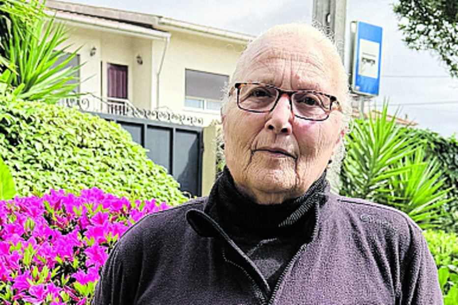 Mulher idosa junto a flores e vegetação, com casa e paragem de autocarro em fundo