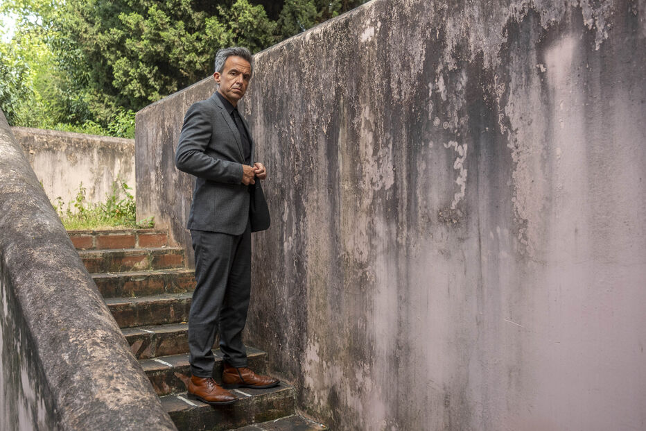 Homem de fato e sapatos castanhos posa nas escadas junto a uma parede