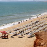 Praia lotada com chapéus de sol, mar e pessoas, com debate sobre concessões