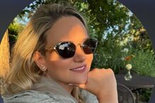 Mulher sorri com óculos de sol e pulseira dourada num ambiente verdejante