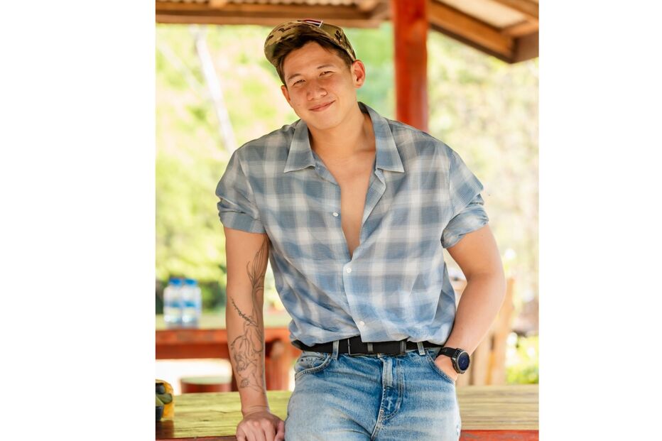 Homem sorridente com camisa axadrezada, calças de ganga e boné camuflado num espaço aberto