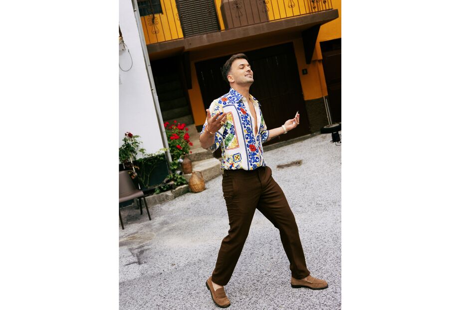 Homem com camisa florida e calças castanhas posa em frente a casas coloridas