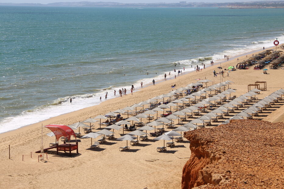 Praia lotada com chapéus de sol, mar e pessoas, com debate sobre concessões