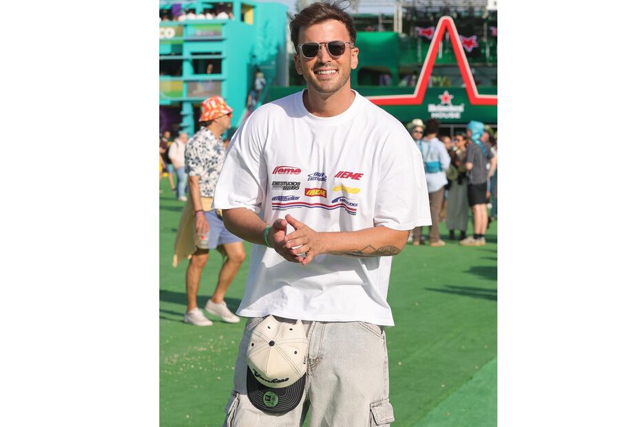 Um homem sorri com óculos de sol e t-shirt com logos, junto a um palco no festival NOS Alive