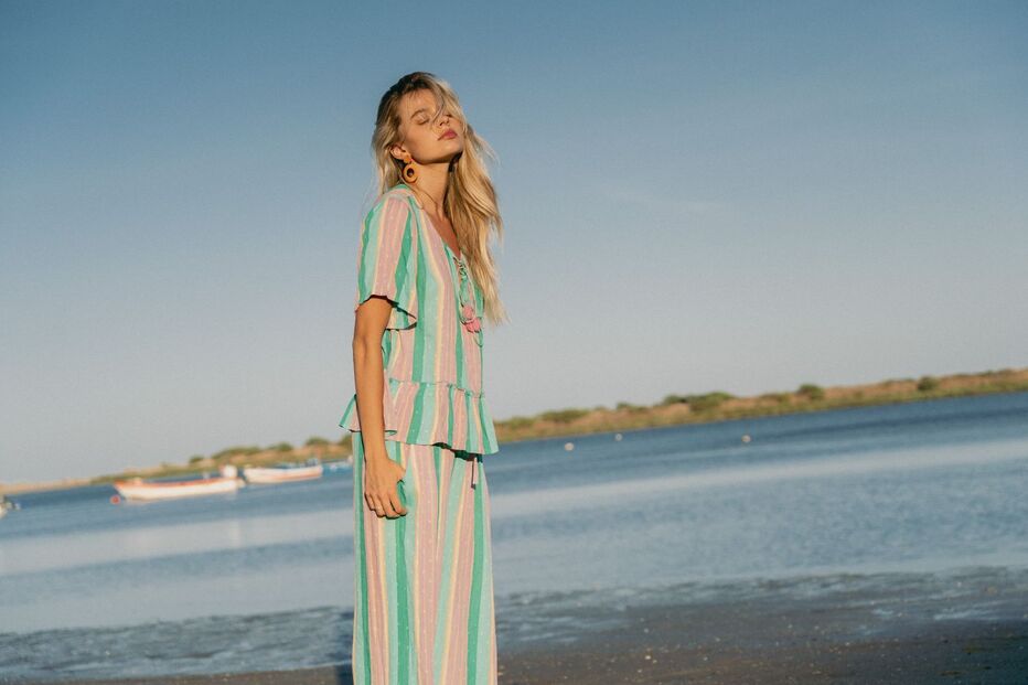 Mulher de cabelo loiro, de olhos fechados, usa um vestido às riscas coloridas numa praia