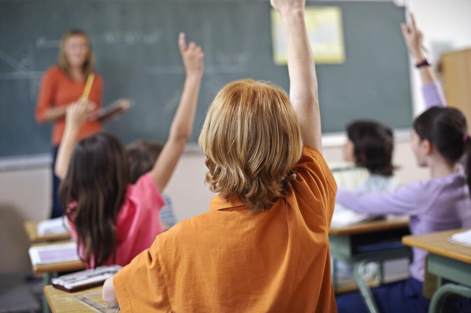 Alunos levantam a mão na sala de aula com a professora ao fundo
